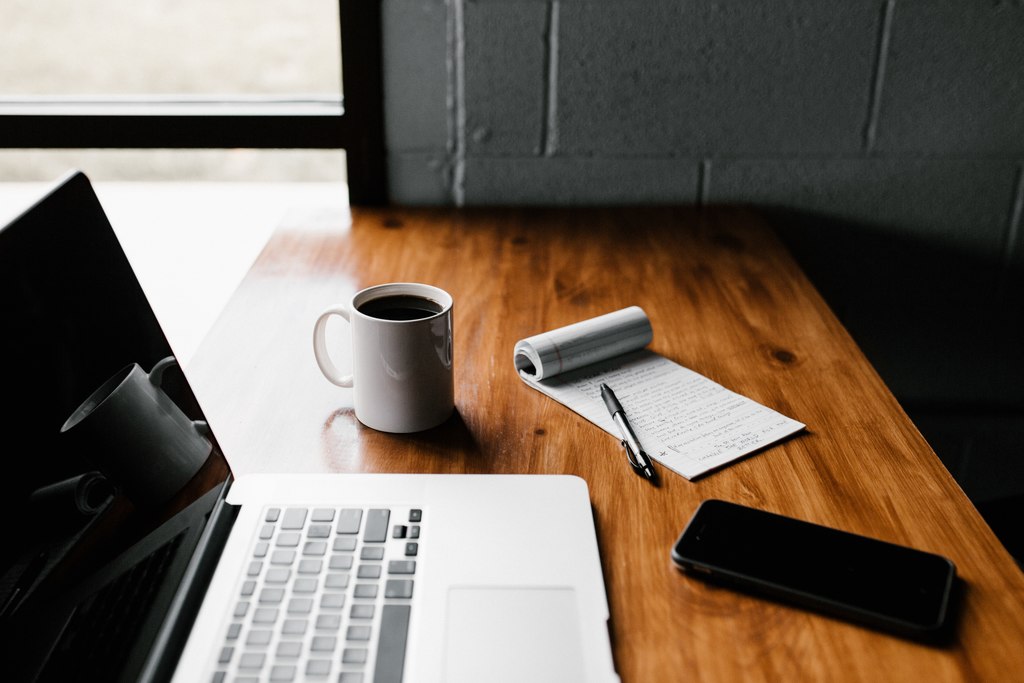 Escrivaninha com notebook, caneca com café, bloco de anotações, caneta e celular. Representando como estudar português para concurso