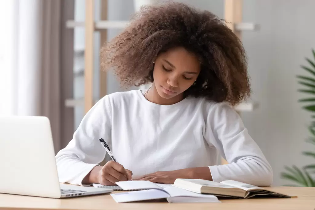 Jovem concentrada em seu caderno, fazendo anotações, enquanto estuda redação para concursos
