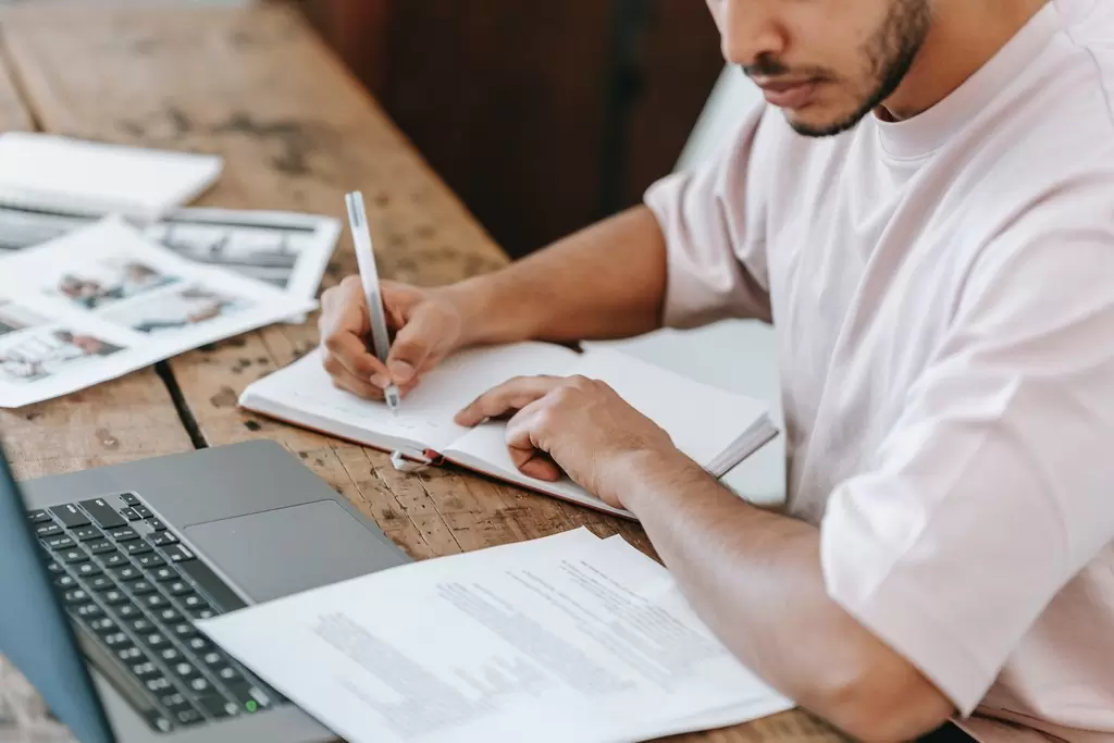 Mão de homem fazendo anotações em um caderno com uma caneta enquanto analisa um edital de concurso público no notebook