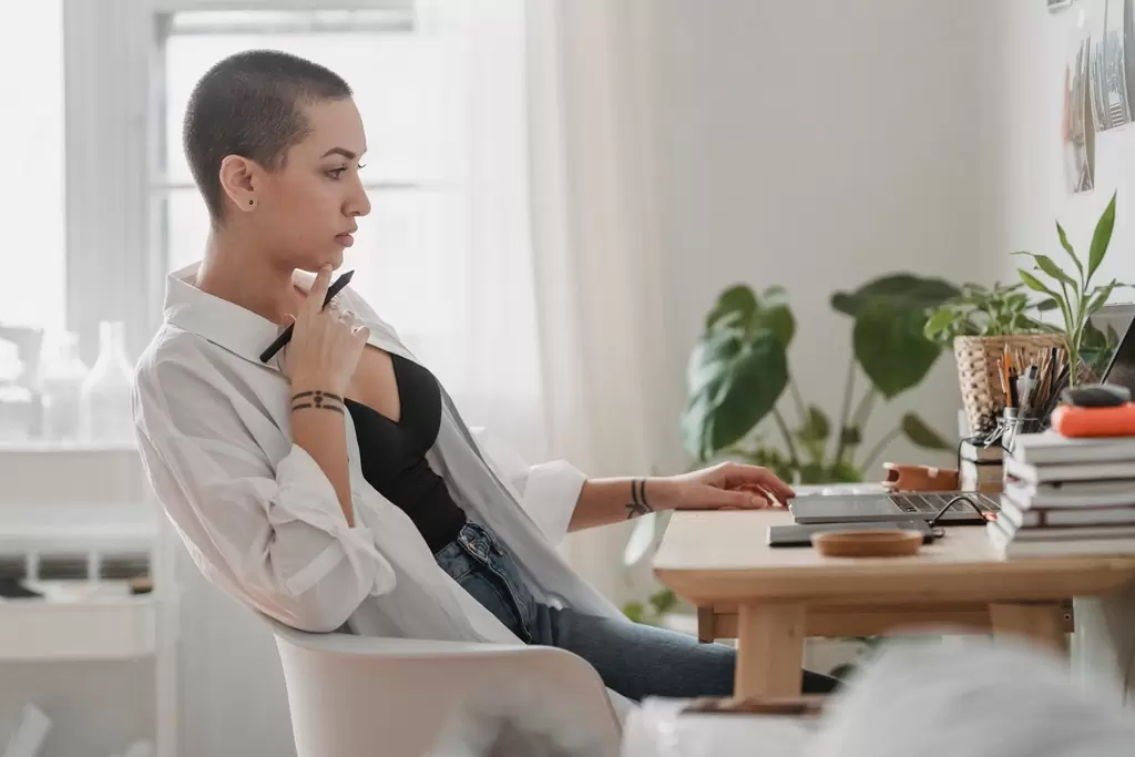 Mulher com dedo no queixo, segurando caneta e olhando concentrada olhando para um notebook enquanto analisa um edital de concurso