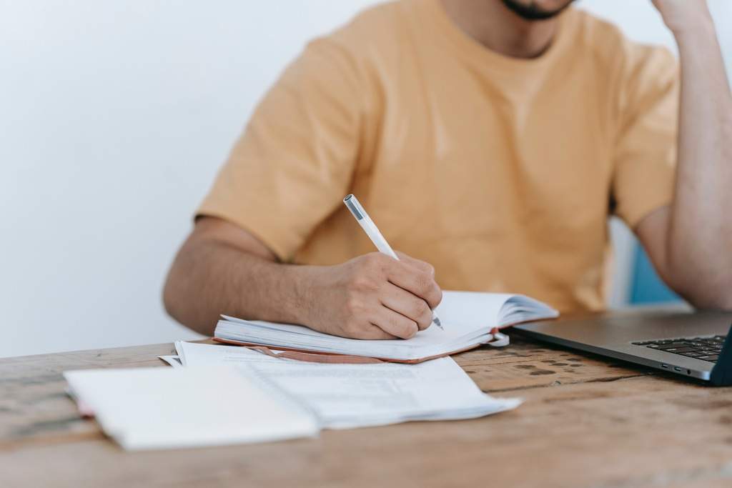 Homem em frente à mesa, segurando caneta, escrevendo em caderno, olhando para notebook, enquanto lê o edital de concurso