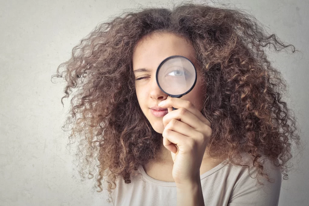 Mulher com blusa cinza segura uma lupa e observa através da lente que aumenta a imagem de seu olho