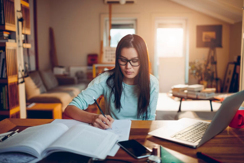 Estudante concentrada escreve em caderno em cima da mesa de estudos enquanto estuda questões de crase no notebook