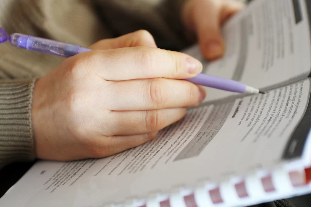 Mãos seguram um lápis lilás e folhas de papel que contém questões de período composto para concursos