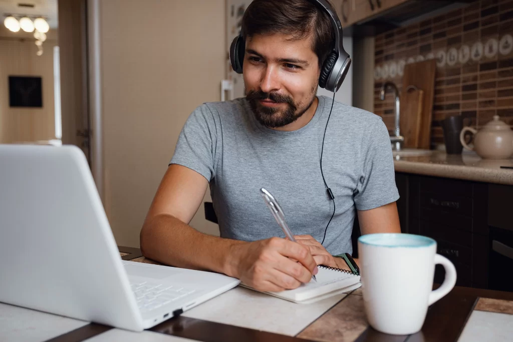 Estudante olha para a tela do notebook e faz anotações em um bloco de notas enquanto estuda com questões de período composto 