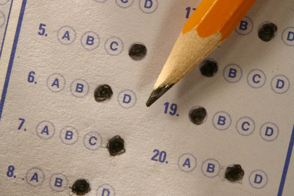 Folha de respostas de concurso público com questões de período composto marcadas por um lápis grafite amarelo