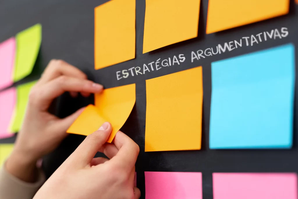 Mãos de aluno colando papéis adesivos coloridos para mapa mental em um quadro negro onde se lê 'estratégias argumentativas'