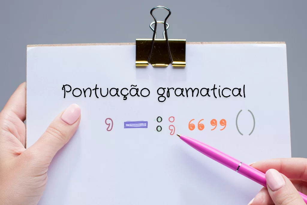 Mão segura uma prancheta com um lápis rosa apontando para os sinais de pontuação gramatical.