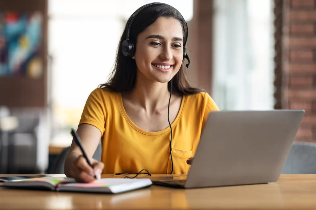 Estudante feliz de blusa amarela fazendo anotações enquanto estuda questões de transitividade verbal para concurso no notebook