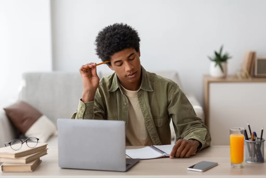 Um concurseiro coça a cabeça com um lápis enquanto examina um caderno à frente de um notebook, onde estão disponíveis simulados gratuitos para concursos