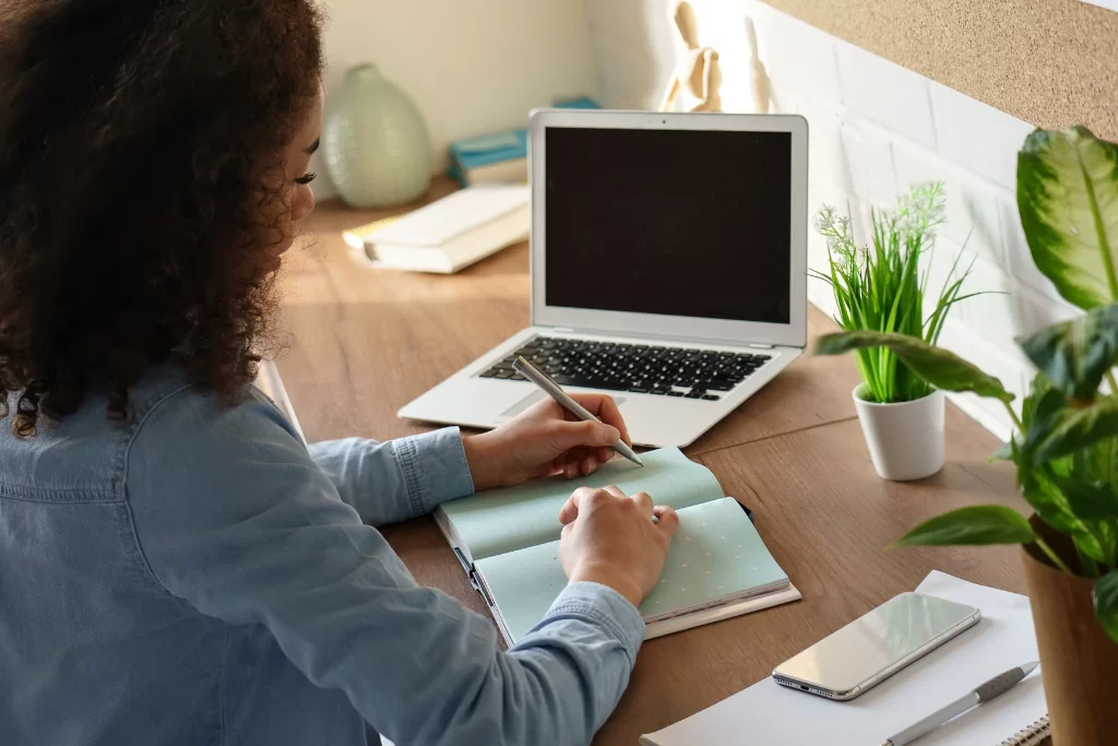 Jovem concentrada em seu caderno estudando tempos, modos e flexões verbais em frente ao notebook
