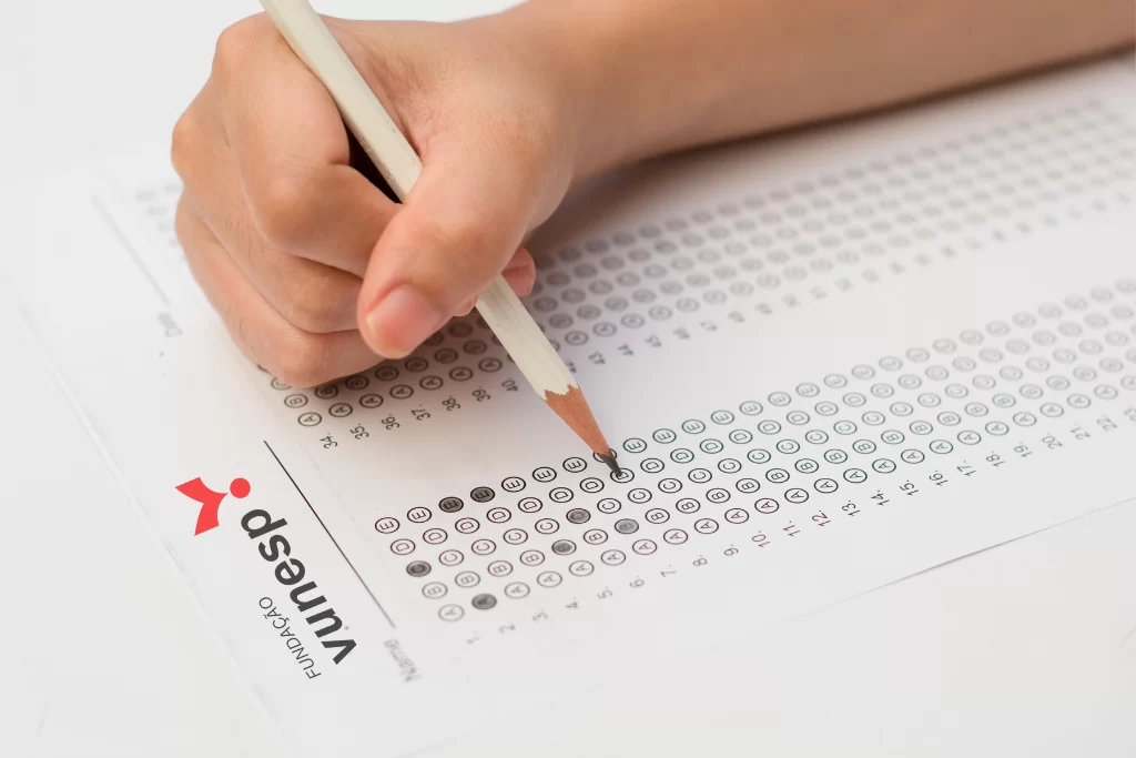 Mão de concurseiro preenchendo o gabarito de prova com modelos de questões da Vunesp