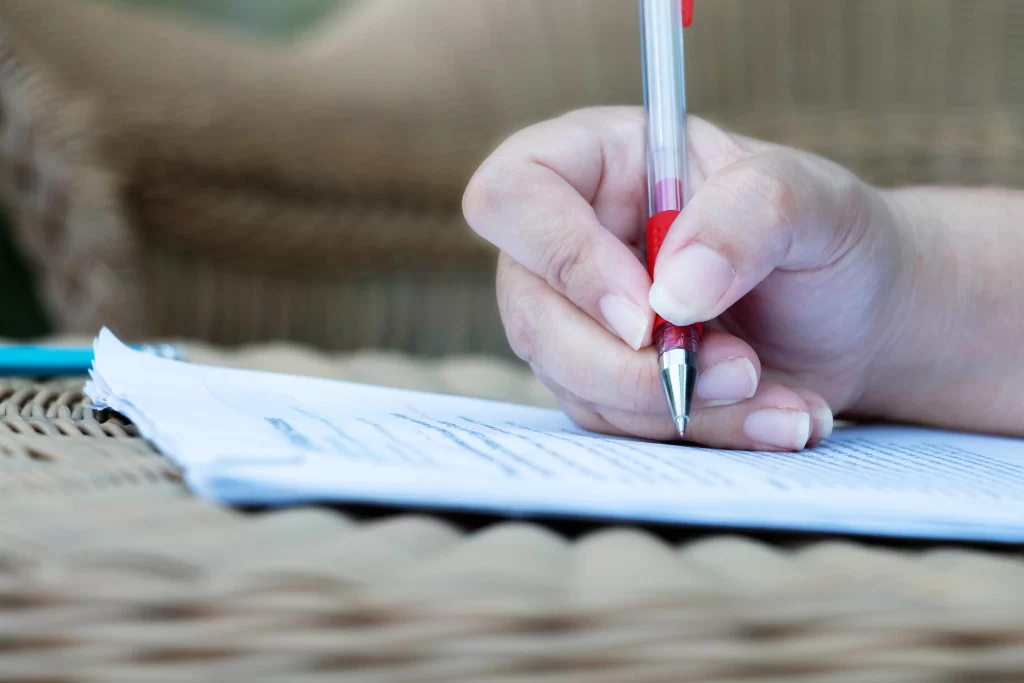 Mão de concurseiro fazendo anotações em um caderno com uma caneta vermelha enquanto estuda 'pronome relativo, o que é?'