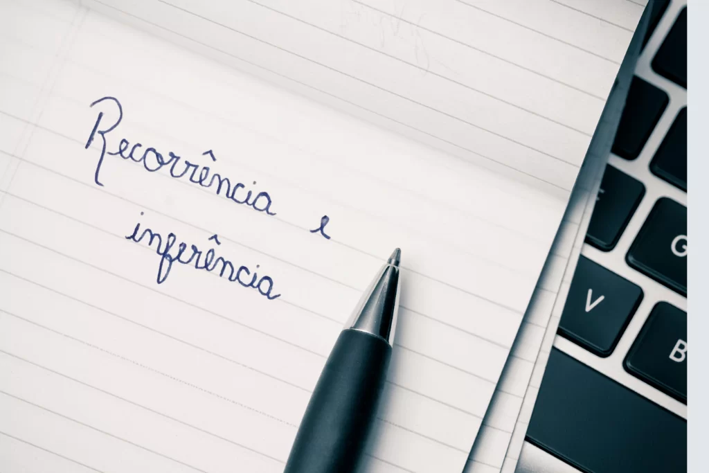 Detalhe de caneta sobre papel pautado com a inscrição 'recorrência e inferência', posicionada sobre um notebook