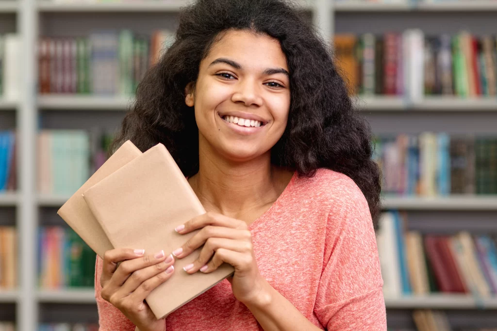 Estudante sorridente segura cadernos de anotações em biblioteca enquanto estuda Português para concursos do zero