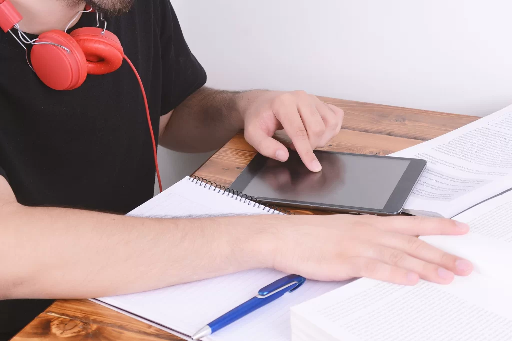 Concurseiro com fones vermelhos estuda sobre modo subjuntivo do verbo em mesa com caneta, apostilas e tablet