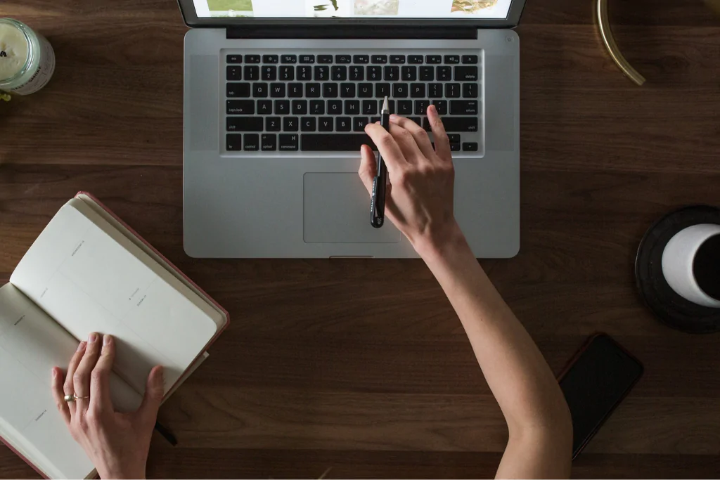 Concurseira segurando um caderno aberto com uma mão e utilizando a outra para mexer no touchpad do notebook, enquanto estuda conjução integrante.