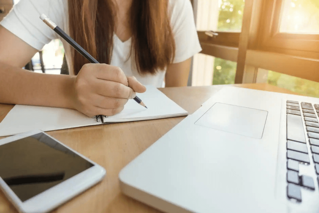 Concurseira de blusa branca escreve em caderno com lápis preto enquanto estuda adjetivo e advérbio