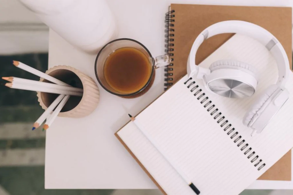 Mesa com caderno aberto, xícara de café, fones de ouvidos e lápis brancos para estudar período simples e composto mapa mental